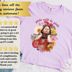 May include: Light purple t-shirt with the phrase "girls will be girls" in pink script. The shirt features a floral design with an image of a person. The shirt has a round neckline.