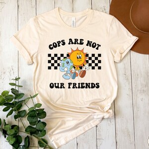 May include: A beige t-shirt with a graphic of a sun and earth holding hands. The graphic is surrounded by a black and white checkered pattern. The text "COPS ARE NOT OUR FRIENDS" is printed in black letters on the shirt.