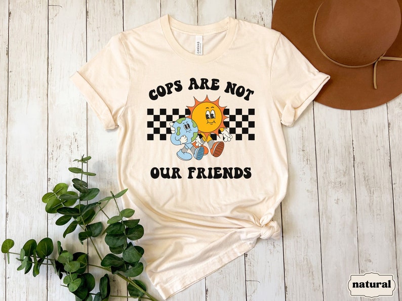 May include: A beige t-shirt with a graphic of a sun and earth holding hands. The graphic is surrounded by a black and white checkered pattern. The text "COPS ARE NOT OUR FRIENDS" is printed in black letters on the shirt.