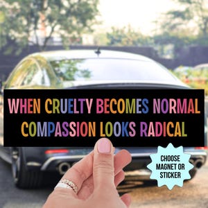 May include: A black rectangular magnet or sticker with the text "WHEN CRUELTY BECOMES NORMAL COMPASSION LOOKS RADICAL" in colorful letters. The background shows a car and a hand holding the magnet.