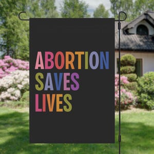 May include: A black garden flag with the text "ABORTION SAVES LIVES" in rainbow colors.