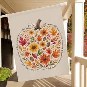 Puede incluir: Una bandera de jardín de color crema con un diseño de calabaza lleno de hojas de otoño, girasoles y bellotas. La calabaza está rodeada por el texto "Fuck Trump". La bandera está colgada de un poste blanco en un porche.