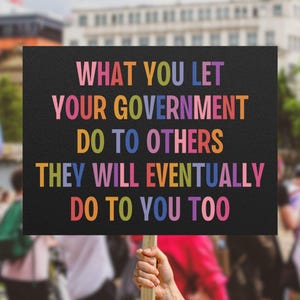 May include: A black sign held up in a crowd with the text "WHAT YOU LET YOUR GOVERNMENT DO TO OTHERS THEY WILL EVENTUALLY DO TO YOU TOO" in colorful letters. A rainbow flag is visible in the background.