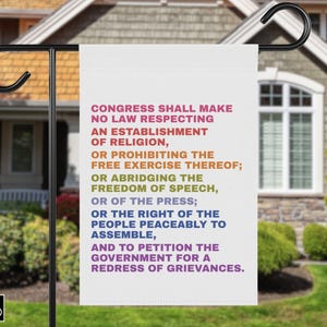 May include: A white garden flag with the text of the First Amendment in various colors. The flag is hanging from a black metal pole in front of a house with a green lawn and landscaping.