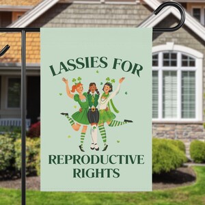May include: A green garden flag with the text "Lassies for Reproductive Rights" and an illustration of three women in green dresses and shamrock hats.