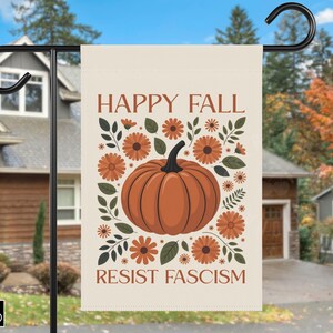 Può includere: Una bandiera da giardino beige con le parole "HAPPY FALL" e "RESIST FASCISM" in lettere marroni. La bandiera presenta una grande zucca arancione circondata da fiori arancioni e foglie verdi. La bandiera è appesa a un palo di metallo nero.