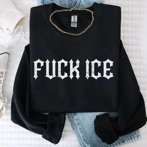 May include: Black sweatshirt with the text "FUCK ICE" in a white gothic font. A gold chain necklace rests on the sweatshirt. White high-top sneakers and denim jeans are visible in the background.