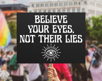 May include: A black sign with the words "BELIEVE YOUR EYES, NOT THEIR LIES" in white, stylized font. Below the text is a white eye symbol with radiating lines. The sign is held up in a crowd, with a rainbow flag visible in the background.