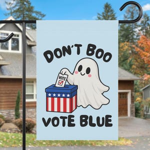Może przedstawiać: Jasnoniebieska flaga ogrodowa z białym duszkiem wrzucającym kartę do głosowania do czerwono-biało-niebieskiej urny. Napis DON'T BOO VOTE BLUE jest wykonany czarnymi literami. Duszek ma różowe policzki, a na karcie widnieje czerwony znaczek.