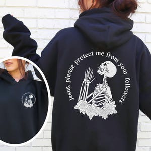 Puede incluir: Sudadera con capucha negra con un gráfico blanco de un esqueleto rezando con el texto "Jesús, por favor, protégeme de tus seguidores".