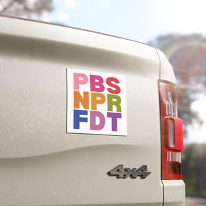 May include: A white rectangular magnet with the text "PBS NPR FDT" in colorful block letters. The magnet is affixed to the back of a white vehicle.
