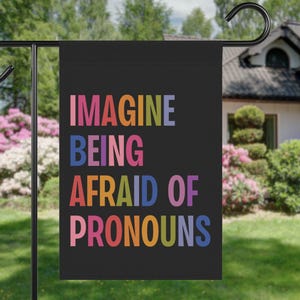 Può includere: Una bandiera da giardino nera con il testo "IMAGINE BEING AFRAID OF PRONOUNS" in colori arcobaleno.