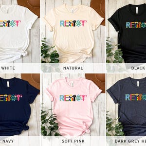 Puede incluir: Seis camisetas de diferentes colores con la palabra "RESIST" escrita en letras de colores. Cada letra est&aacute; decorada con un s&iacute;mbolo diferente, incluyendo un pu&ntilde;o, un &uacute;tero, un &aacute;tomo y un arco&iacute;ris. Las camisetas se muestran sobre un fondo de madera.