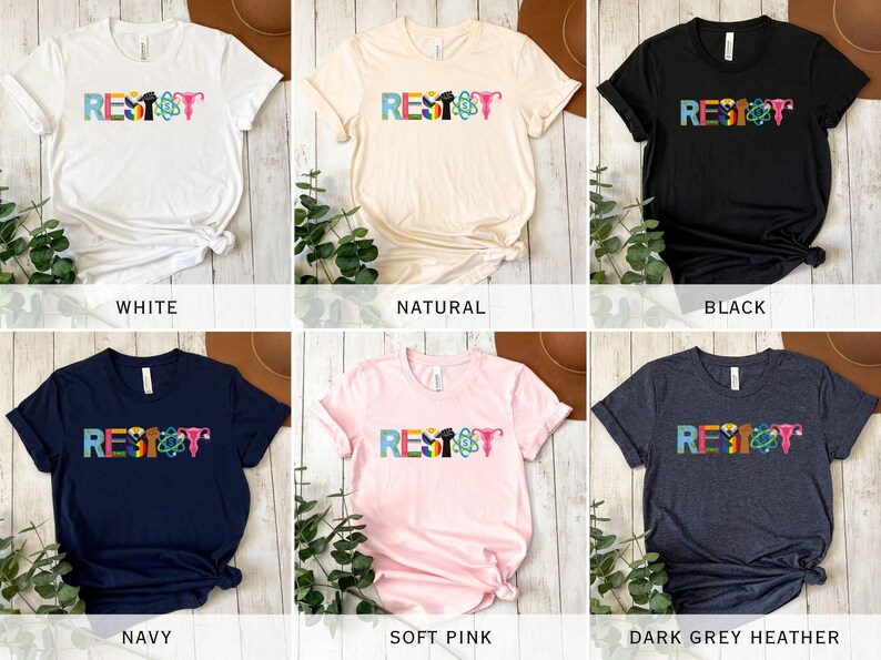 Puede incluir: Seis camisetas de diferentes colores con la palabra "RESIST" escrita en letras de colores. Cada letra est&aacute; decorada con un s&iacute;mbolo diferente, incluyendo un pu&ntilde;o, un &uacute;tero, un &aacute;tomo y un arco&iacute;ris. Las camisetas se muestran sobre un fondo de madera.