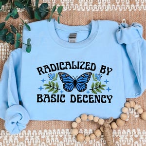 Puede incluir: Sudadera azul claro con el texto "RADICALIZED BY BASIC DECENCY" en negro. Un gráfico de mariposa azul está centrado sobre el texto, con flores azules y hojas verdes.