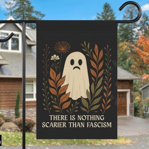 May include: Black garden flag with a sad ghost illustration and botanical details in brown, green, and cream. The flag reads "THERE IS NOTHING SCARIER THAN FASCISM". Decorative outdoor Halloween decor.