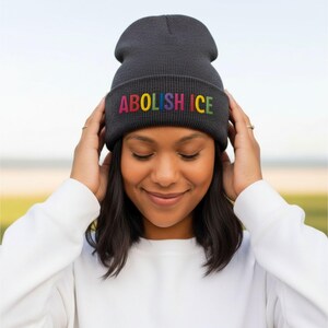 May include: A dark gray beanie with the embroidered text "ABOLISH ICE" in rainbow colors. The beanie is being held by a person wearing a white long-sleeved shirt. The background is blurred, with a focus on the beanie.