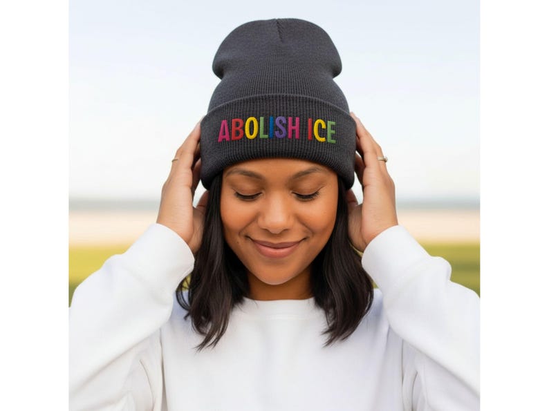 May include: A dark gray beanie with the embroidered text "ABOLISH ICE" in rainbow colors. The beanie is being held by a person wearing a white long-sleeved shirt. The background is blurred, with a focus on the beanie.