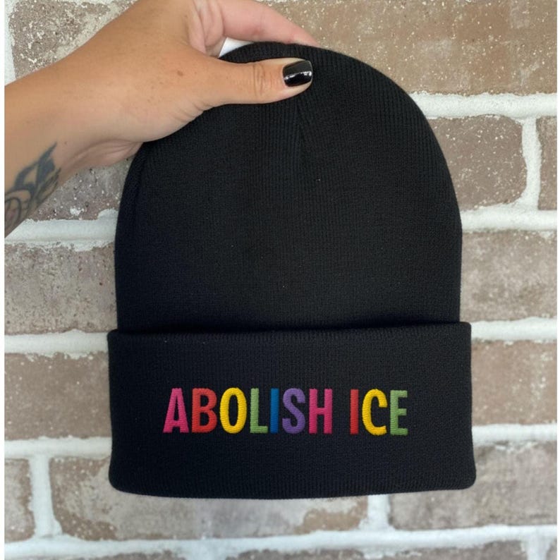 May include: Black beanie with the embroidered text "ABOLISH ICE" in rainbow colors. The beanie is made of a soft, knitted material and has a folded brim. The text is centered on the brim.