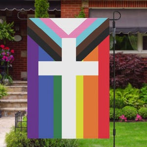 Puede incluir: Una bandera de jardín con una cruz blanca sobre un fondo de rayas de arcoíris, que incluyen morado, azul, verde, amarillo, naranja y rojo. La cruz se superpone a un triángulo marrón, azul y rosa.