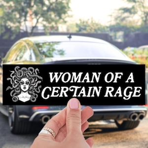 May include: Black rectangular bumper sticker with the text "WOMAN OF A CERTAIN RAGE" in white. The sticker features a black and white illustration of Medusa wearing sunglasses. The sticker is held in front of a car.