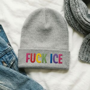 May include: A gray beanie with the embroidered text "FUCK ICE" in rainbow colors. The beanie is folded at the brim and is displayed with a denim jacket and a gray scarf.