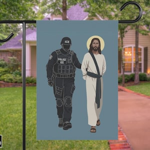 May include: A garden flag depicting Jesus being escorted by an ICE officer. The officer is in a black uniform with the text "POLICE ICE". Jesus is in white robes with a halo. The flag is on a black metal stand.