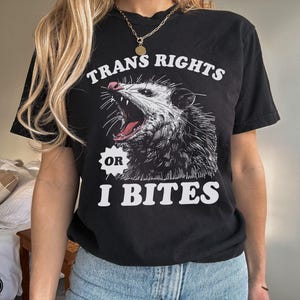 Puede incluir: Camiseta negra con un gráfico blanco de un opossum con el texto "Trans Rights or I Bites".