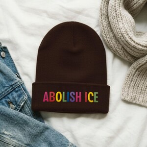 May include: A brown beanie with the embroidered text "ABOLISH ICE" in rainbow colors. The hat is made of a soft material and is folded at the brim. The beanie is displayed on a white surface with a denim jacket and a gray scarf.