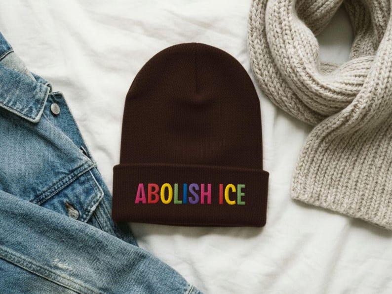 May include: A brown beanie with the embroidered text "ABOLISH ICE" in rainbow colors. The hat is made of a soft material and is folded at the brim. The beanie is displayed on a white surface with a denim jacket and a gray scarf.