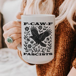 May include: A white ceramic mug featuring a black graphic illustration of a crow with wings spread, surrounded by floral vines. The text F-CAW-F is printed above the bird, and the word FASCISTS is printed below the bird in a bold black font.