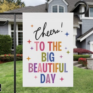 May include: A white garden flag with the words "Cheers! TO THE BIG BEAUTIFUL DAY" in colorful text. The flag is decorated with star shapes and is displayed on a black metal stand in a yard.