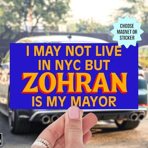 May include: A blue rectangular magnet or sticker with the text "I MAY NOT LIVE IN NYC BUT ZOHRAN IS MY MAYOR" in yellow and orange. The background shows a car and a hand holding the magnet.