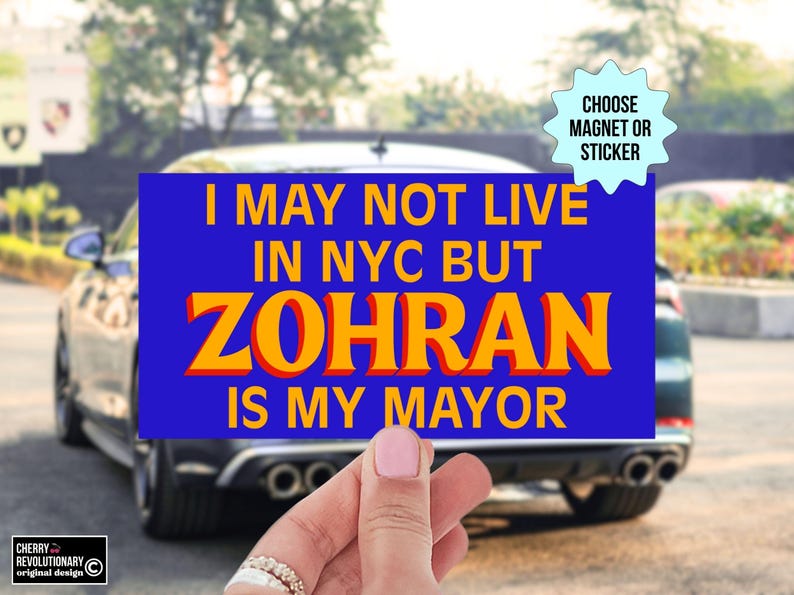 May include: A blue rectangular magnet or sticker with the text "I MAY NOT LIVE IN NYC BUT ZOHRAN IS MY MAYOR" in yellow and orange. The background shows a car and a hand holding the magnet.