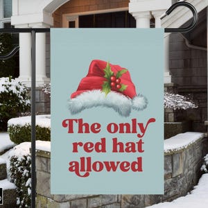 Könnte beinhalten: Eine Gartenflagge mit hellblauem Hintergrund zeigt einen roten Weihnachtsmannhut mit weißem Besatz und Stechbeeren. Der Text "The only red hat allowed" ist in roter Schrift. Die Flagge wird im Freien gezeigt.