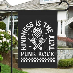 May include: Black garden flag with white text that reads "KINDNESS IS THE REAL PUNK ROCK." The design includes crossed skeleton hands, a checkered heart, and flames. The flag is displayed outdoors.