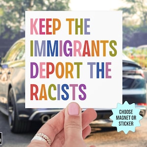 May include: A white square magnet or sticker with the text "KEEP THE IMMIGRANTS DEPORT THE RACISTS" in colorful block letters. The image is held in front of a car.