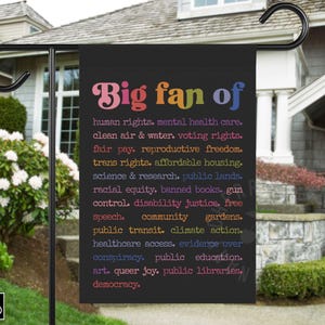 Könnte beinhalten: Schwarze Gartenflagge mit dem Text "Big fan of" in bunten Buchstaben, gefolgt von einer Liste von Menschenrechten und sozialen Gerechtigkeitsfragen. Die Flagge hängt an einem schwarzen Metallständer vor einem Haus.