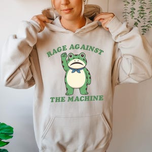 May include: Beige hoodie with a front graphic of a green frog with a raised fist and a blue bow tie. The text "RAGE AGAINST THE MACHINE" is printed above and below the frog in green.