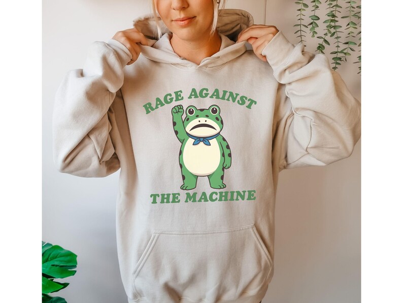May include: Beige hoodie with a front graphic of a green frog with a raised fist and a blue bow tie. The text "RAGE AGAINST THE MACHINE" is printed above and below the frog in green.