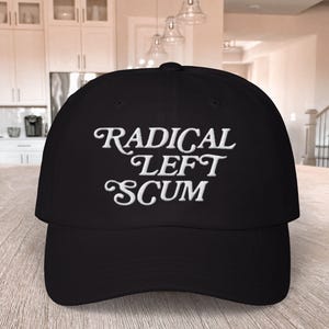 May include: Black baseball cap with the embroidered text "RADICAL LEFT SCUM" in white script. The cap has a curved brim and a structured crown. The hat is displayed on a wooden surface.