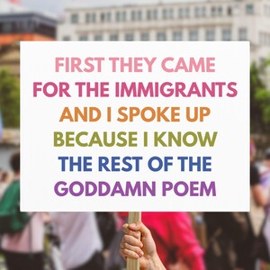 May include: A white sign with the text "FIRST THEY CAME FOR THE IMMIGRANTS AND I SPOKE UP BECAUSE I KNOW THE REST OF THE GODDAMN POEM" in rainbow colors. The sign is held up in a crowd, with a rainbow flag visible.
