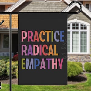 May include: A black garden flag displaying the words "PRACTICE RADICAL EMPATHY" in vibrant, multi-coloured block letters. The flag is suspended on a black metal pole, set against a backdrop of a house and garden.