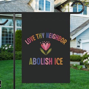 May include: Black garden flag with the text "LOVE THY NEIGHBOR ABOLISH ICE" in colorful letters. A floral design is centered between the phrases. The flag is displayed on a black metal stand in a grassy yard.