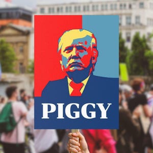 May include: A protest sign with a portrait of a man with yellow hair, red tie, and blue suit, against a red and blue backdrop. The word "PIGGY" is written in white block letters below the portrait.