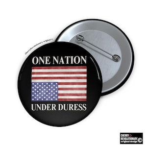 May include: A black button with a white background and the text "ONE NATION UNDER DURESS". The button features a distressed American flag with the stars on the left side of the flag.