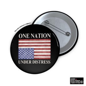 May include: A black button with a white background and the text "ONE NATION UNDER DISTRESS". The button features a distressed American flag with the stars on the wrong side.