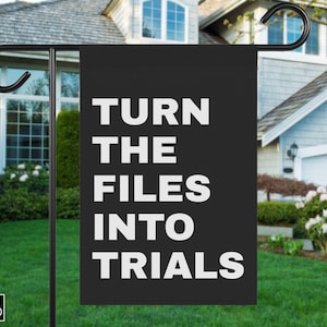 May include: A black garden flag with white text that reads "TURN THE FILES INTO TRIALS." The flag is hanging from a black metal pole in front of a house with a green lawn and bushes.