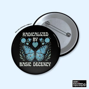 Może przedstawiać: Czarny guzik z napisem "RADICALIZED BY BASIC DECENCY" i niebieskim motylem. Motyl otoczony jest elementami kwiatowymi. Drugi guzik pokazuje tył szpilki.
