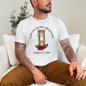 Puede incluir: Camiseta blanca con una ilustraci&oacute;n en blanco y negro de una guillotina con rosas rojas y el texto "Faire les coupes necessaires - Depuis 1789 - "
